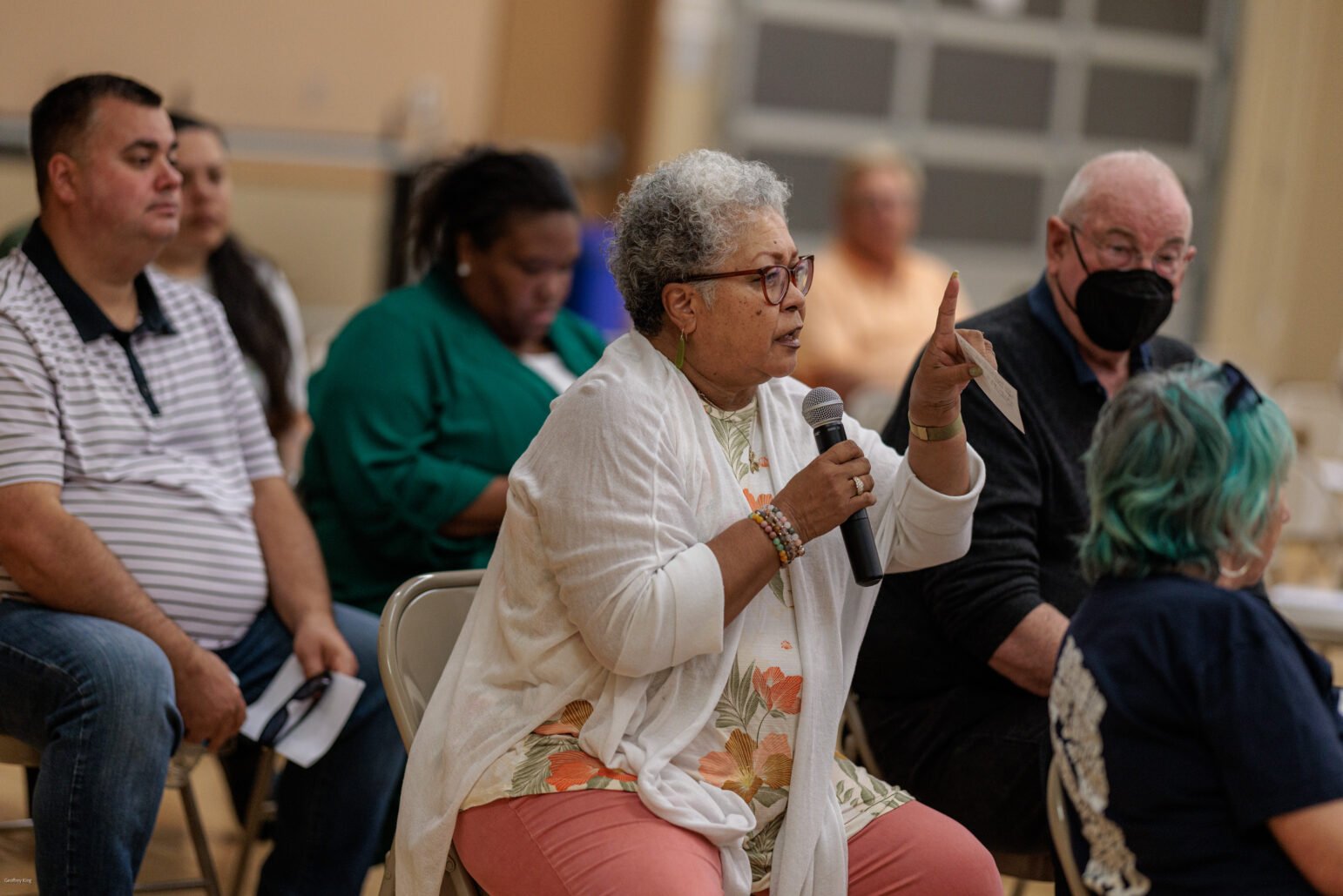 A focused woman with curly gray hair and glasses is seated, speaking into a microphone at a community meeting. She gestures with her hand, indicating a point of discussion. In the background, other attendees listen, with one man wearing a black respiratory mask looking towards her, indicating an engaged audience in a public forum.
