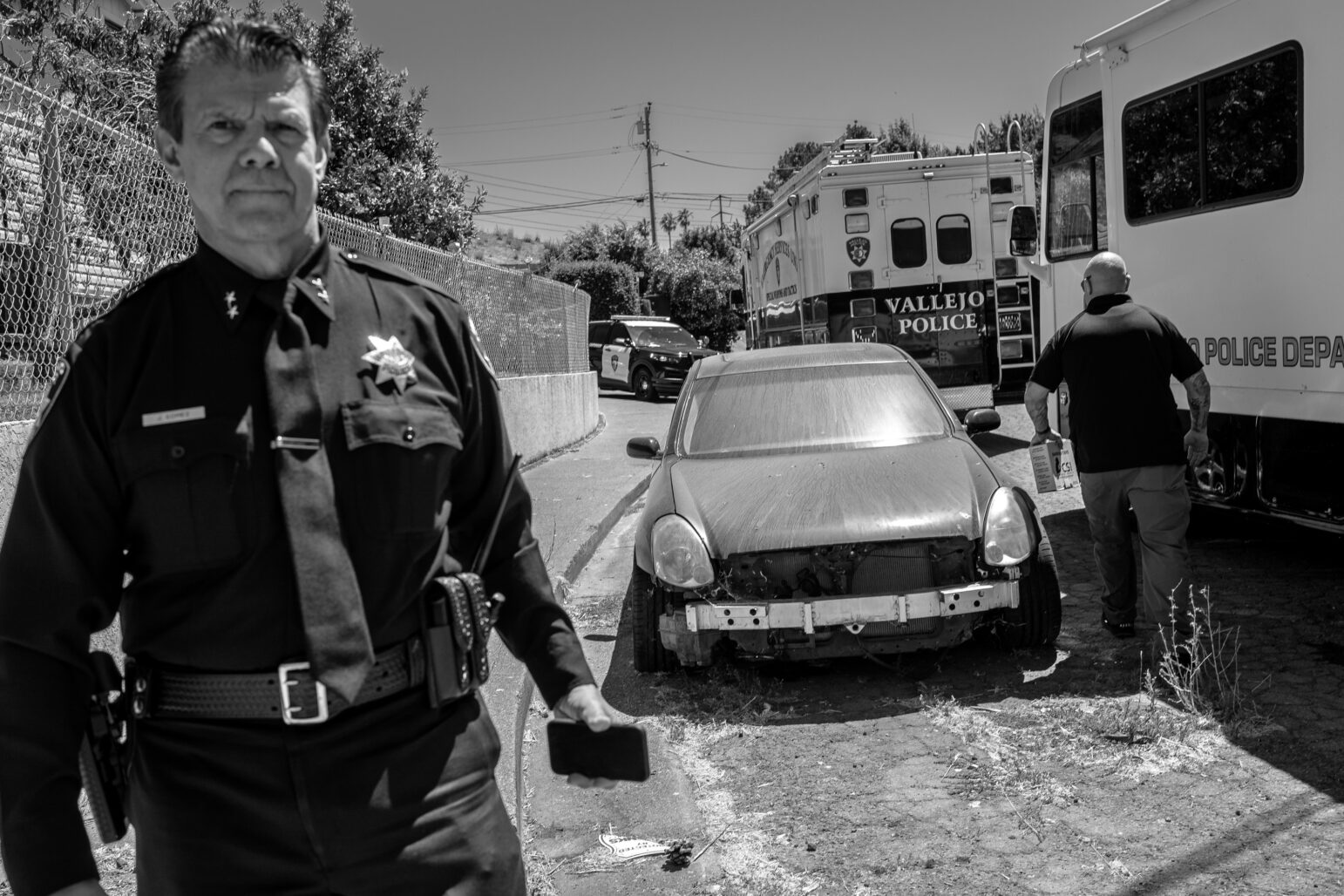 A monochrome image capturing a serious-looking man in a police uniform standing outside on a sunny day. His badge and name tag are visible, and he appears to be holding a cellphone in his left hand. In the background, a dilapidated car is in front of an ambulance and a police department van. There are other indistinct figures and urban details in the scene, with a chain-link fence to the left and power lines above.