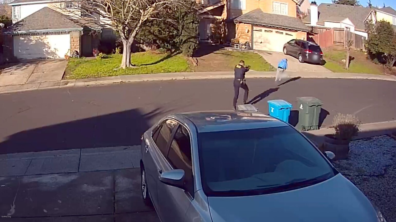 The image, a still from a security camera, shows a suburban street with two individuals and two cars visible. One, a police officer, is in the process of firing a handgun. The second person, dressed in a blue top and dark pants, is falling while in the process of running away toward a house.