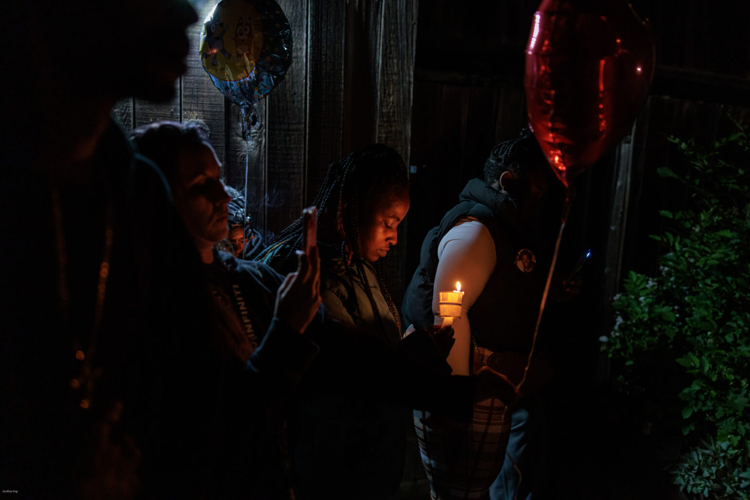 A somber night-time vigil with people holding candles and balloons. The mood is reflective and mournful, with individuals paying respects at a memorial, their faces illuminated softly by the candlelight.