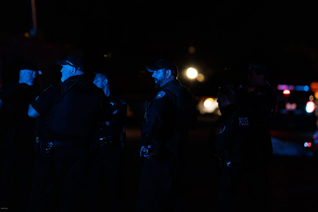 A night scene with a group of police officers gathered in discussion, illuminated by ambient blue and red police car lights in the background, creating a somber atmosphere reflective of a homicide scene investigation.
