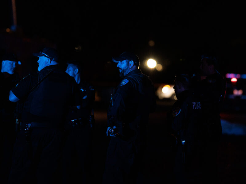 A night scene with a group of police officers gathered in discussion, illuminated by ambient blue and red police car lights in the background, creating a somber atmosphere reflective of a homicide scene investigation.