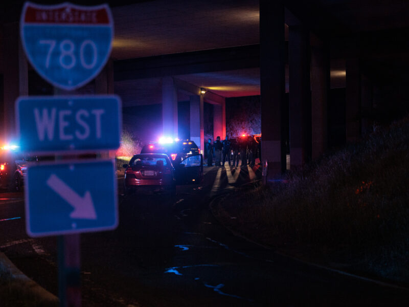 A nighttime shot under an overpass where multiple police officers are gathered near police vehicles with flashing lights, near a road sign indicating "INTERSTATE 780 WEST."