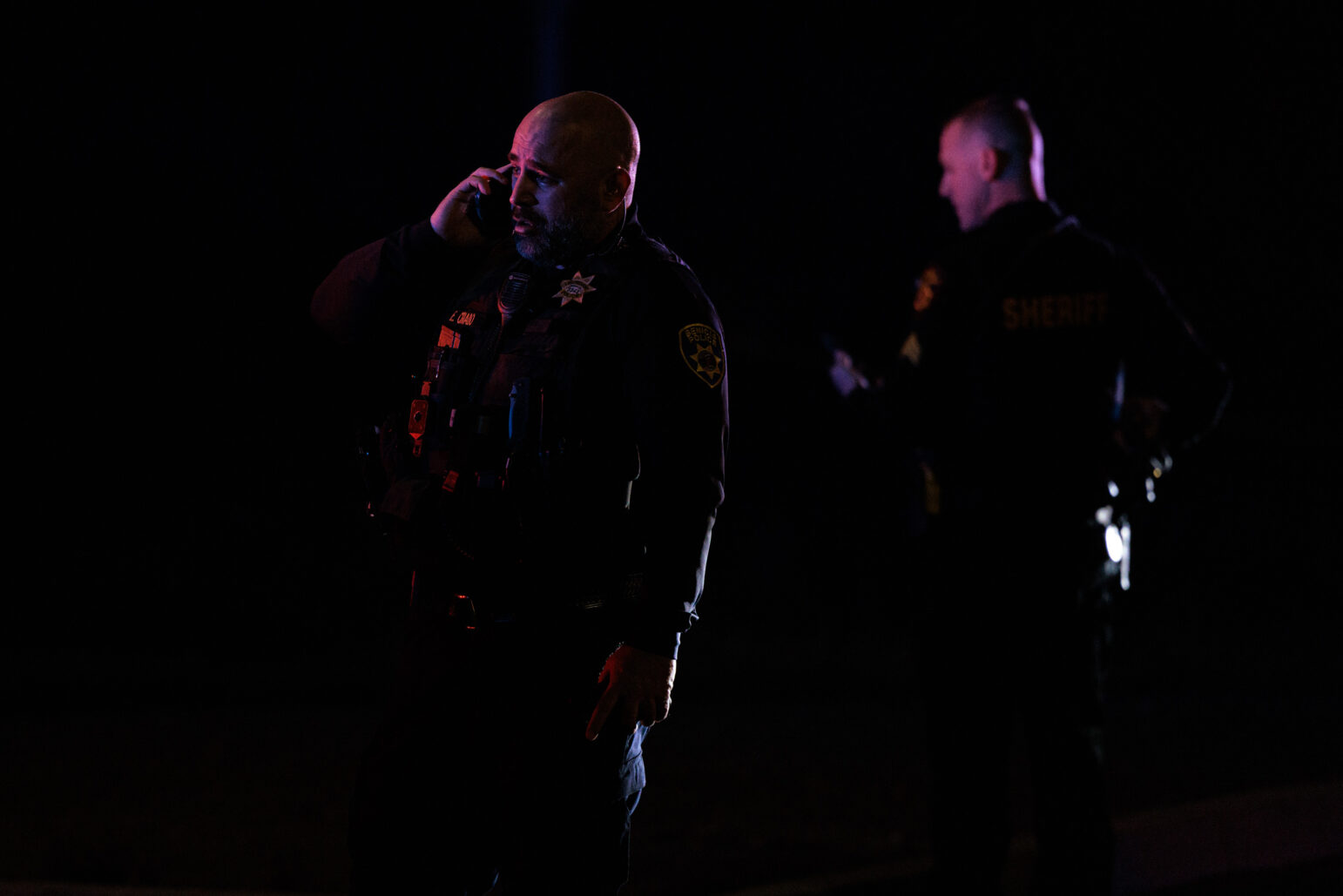 A Benicia police officer speaks on a mobile phone, his face lit by the emergency lights, with another officer in the background. Both are wearing dark uniforms with badges or patches visible.