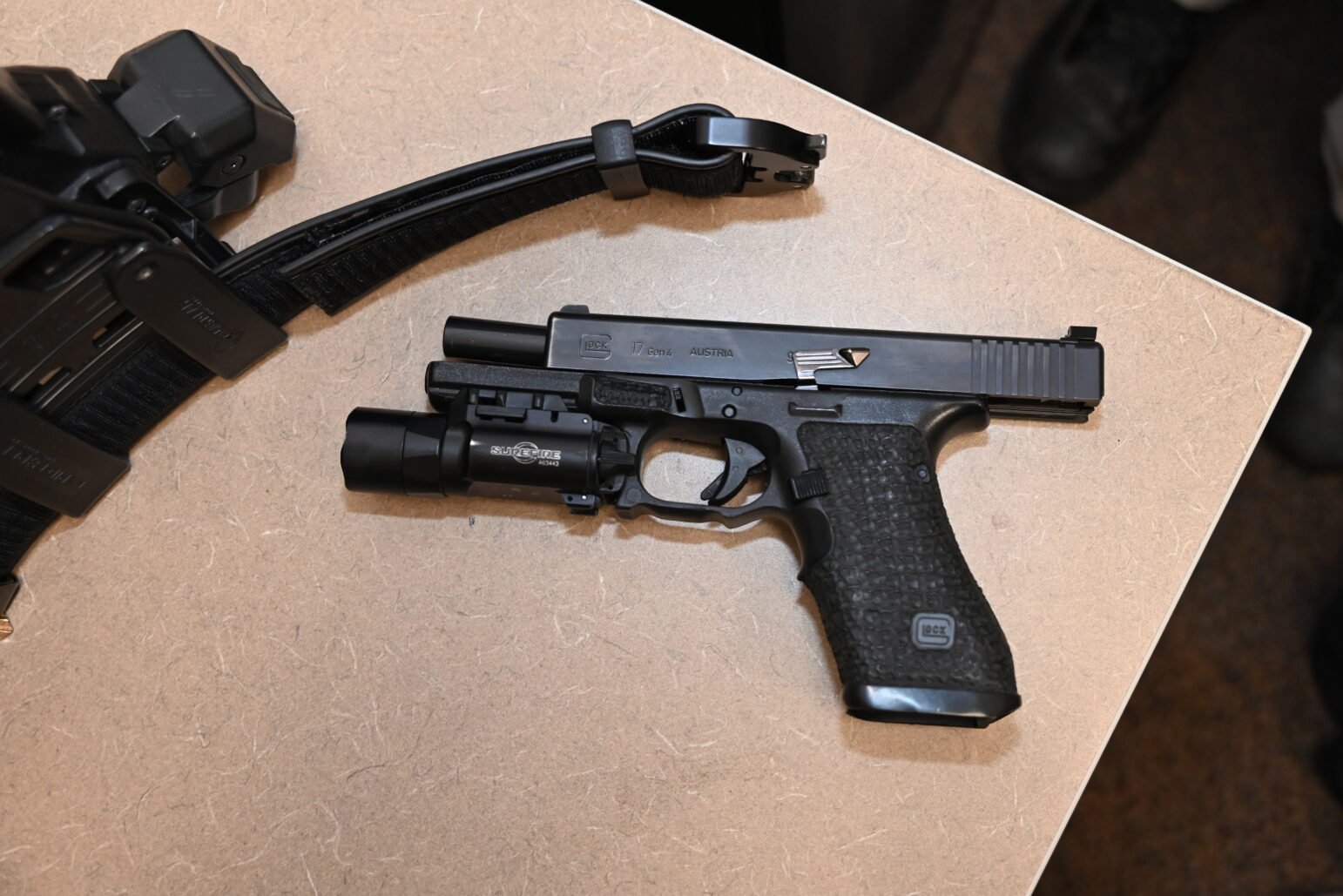 A semi-automatic pistol with a tactical flashlight attached lies on a flat surface alongside a duty belt equipped with various police equipment. The gun and belt are being displayed for documentation or examination, with the pistol's magazine removed and placed next to it.