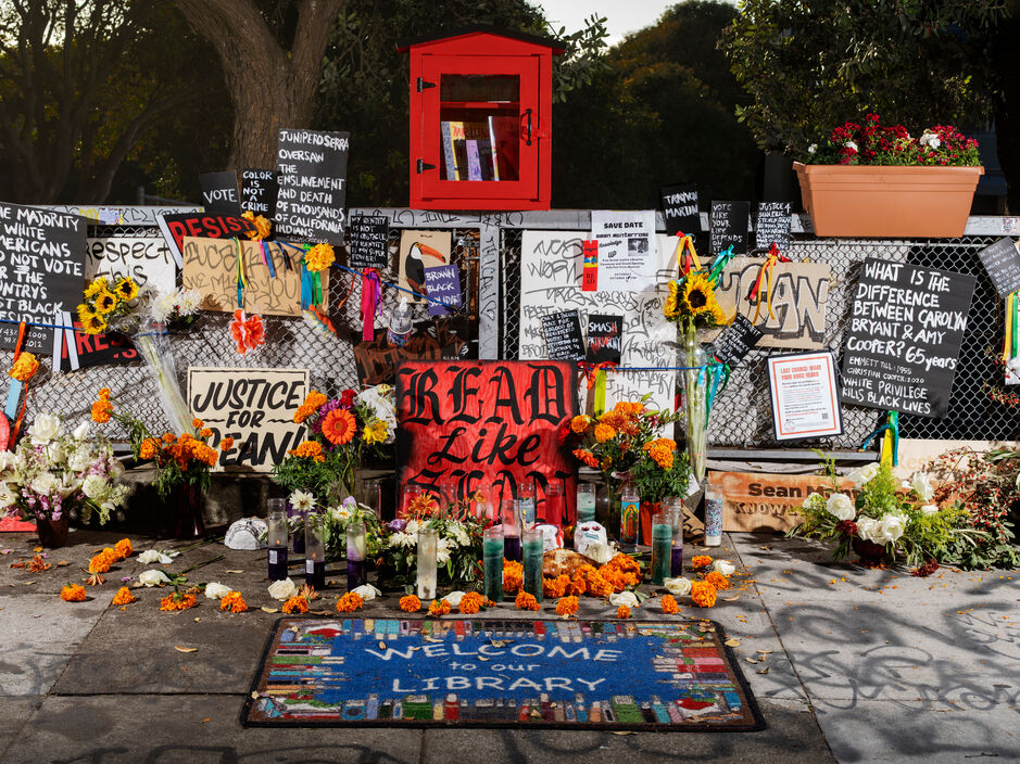 A makeshift memorial with messages, flowers, and candles. The focal point is a colorful altar with handwritten signs, marigolds, sunflowers, and various personal items. One prominent sign reads 'Justice for Sean,' and others contain political and social justice messages. A hand-painted sign on the ground welcomes visitors to a 'Library,' hinting at community solidarity and remembrance.