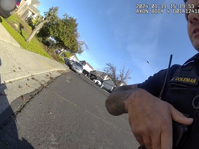 A first-person view from a police officer's body camera, displaying part of the officer's uniform with a name tag reading 'COLEMAN' and a radio speaker microphone attached to the front. The officer's hand is visible, pointing to something off-camera. In the background, a residential street with parked cars and houses can be seen. The sky is clear and blue. Timestamp and camera information are overlaid at the top, showing the date as "2024-01-11 15:19:53 -0800" and the text "AXON BODY 4 D01A12418."