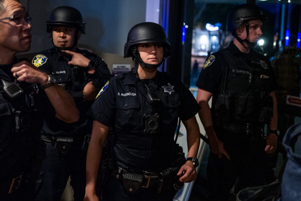 Photo of several police officers in riot gear, standing inside a building. One female officer in the center, identified by a badge as S. McDonough, looks intently ahead. The officers are wearing dark uniforms with helmets, badges, and have body cameras and various equipment attached to their belts.