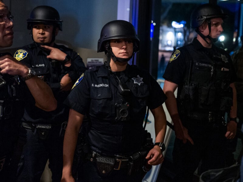Photo of several police officers in riot gear, standing inside a building. One female officer in the center, identified by a badge as S. McDonough, looks intently ahead. The officers are wearing dark uniforms with helmets, badges, and have body cameras and various equipment attached to their belts.