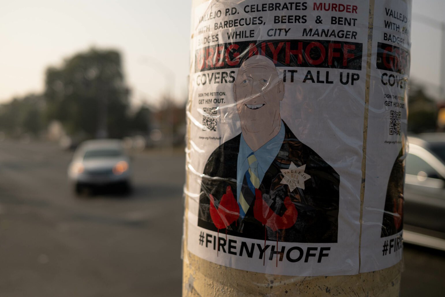 A poster on a utility pole, with a cartoon illustration of a man in a suit, overlaid with text and hashtags calling for his removal from office. There is a QR code at the bottom, and the background shows a blurred street scene.