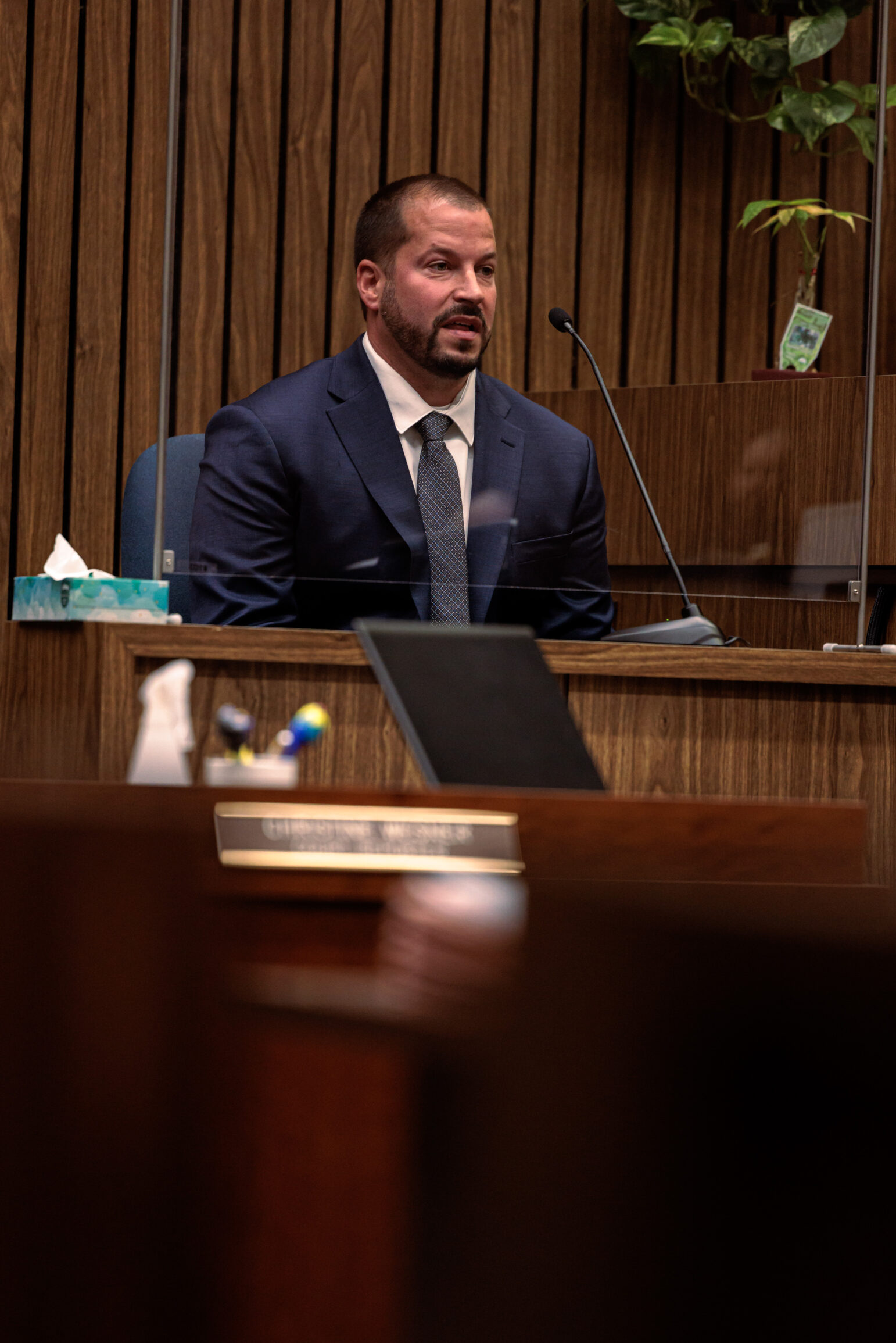 A photo of a man in a business suit sitting in a witness box while giving testimony. He has short hair, a trimmed beard, and wears a blue suit with a tie.