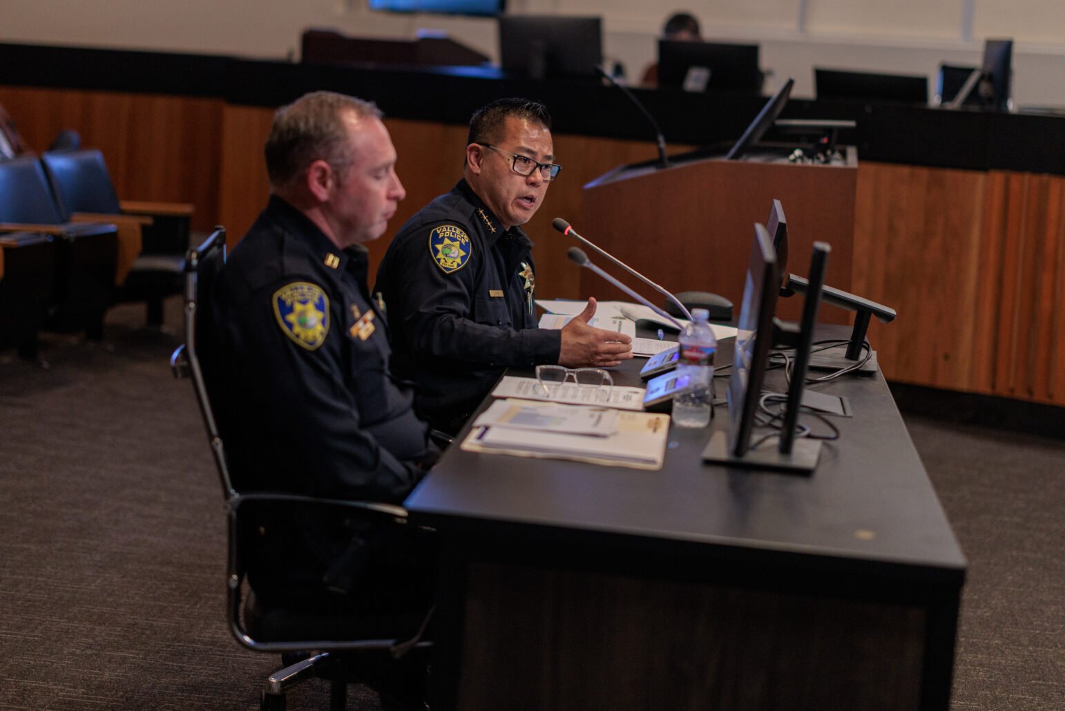 Two police officers are seated at a desk in a council chamber, addressing a public meeting. The officer speaking is wearing glasses and gesturing with his hands, with a microphone in front of him, while his colleague looks on. They are in uniform with badges visible.