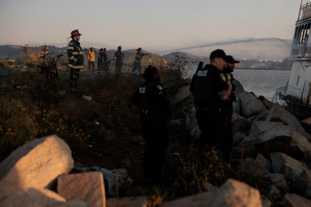 Emergency responders at a waterfront scene during dusk. Police officers and firefighters are scattered across a rocky shoreline with a large vessel visible in the background. Foreground shows rough terrain and some debris, suggesting a recent incident. The sky is orange-hued, indicating either a sunset, and hills are faintly visible in the distance through a light mist.
