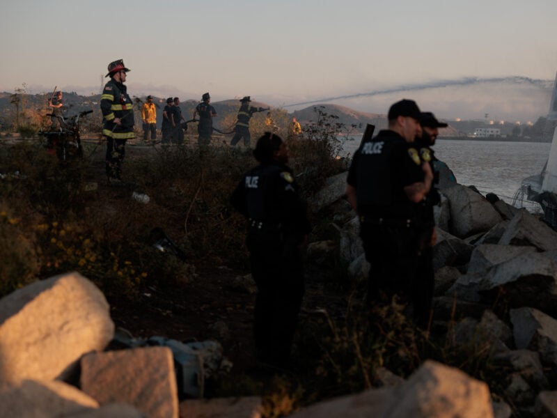 Emergency responders at a waterfront scene during dusk. Police officers and firefighters are scattered across a rocky shoreline with a large vessel visible in the background. Foreground shows rough terrain and some debris, suggesting a recent incident. The sky is orange-hued, indicating either a sunset, and hills are faintly visible in the distance through a light mist.