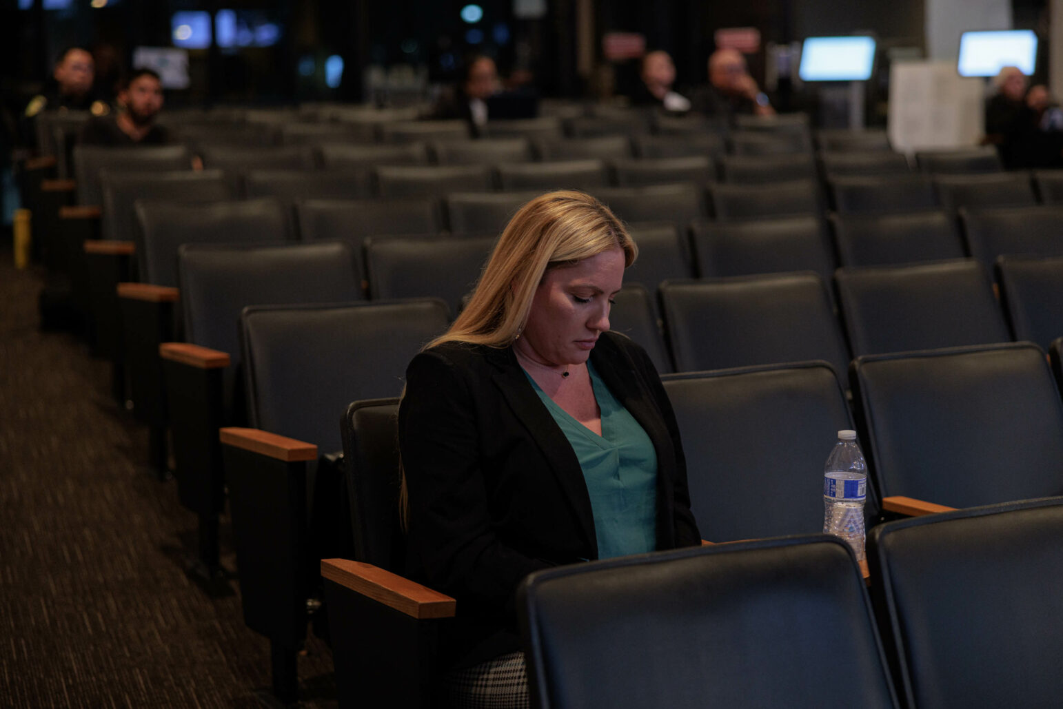A lone woman sits in a large auditorium with rows of empty seats around her. She is looking down at her phone, engrossed in its content. The setting suggests an atmosphere of isolation or reflection amid a public space.