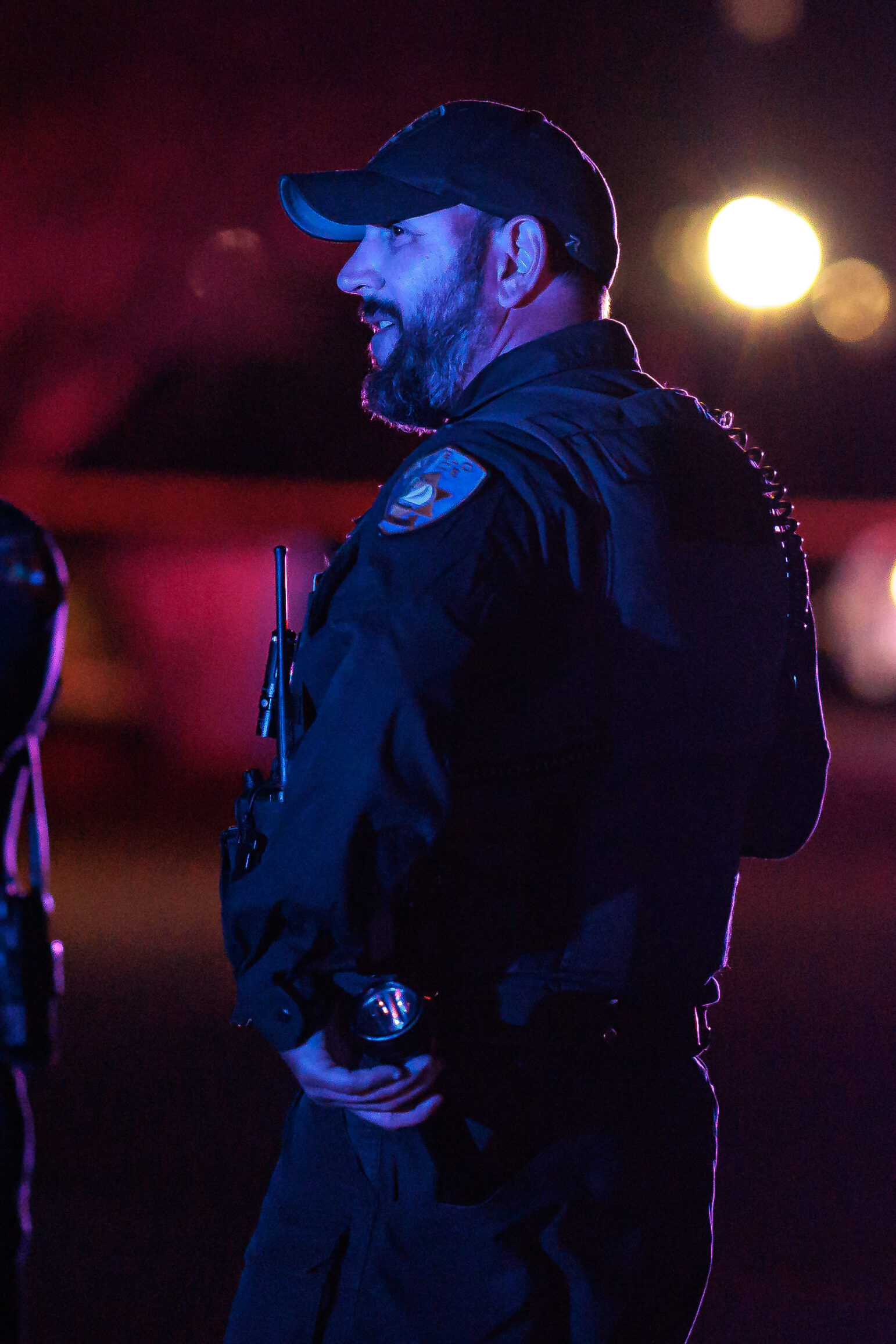 Photo of a police officer standing outdoors at night. The officer is in profile, looking to the left, with a beard and wearing a dark uniform with a badge and a radio. Red and blue police lights softly illuminate the background.