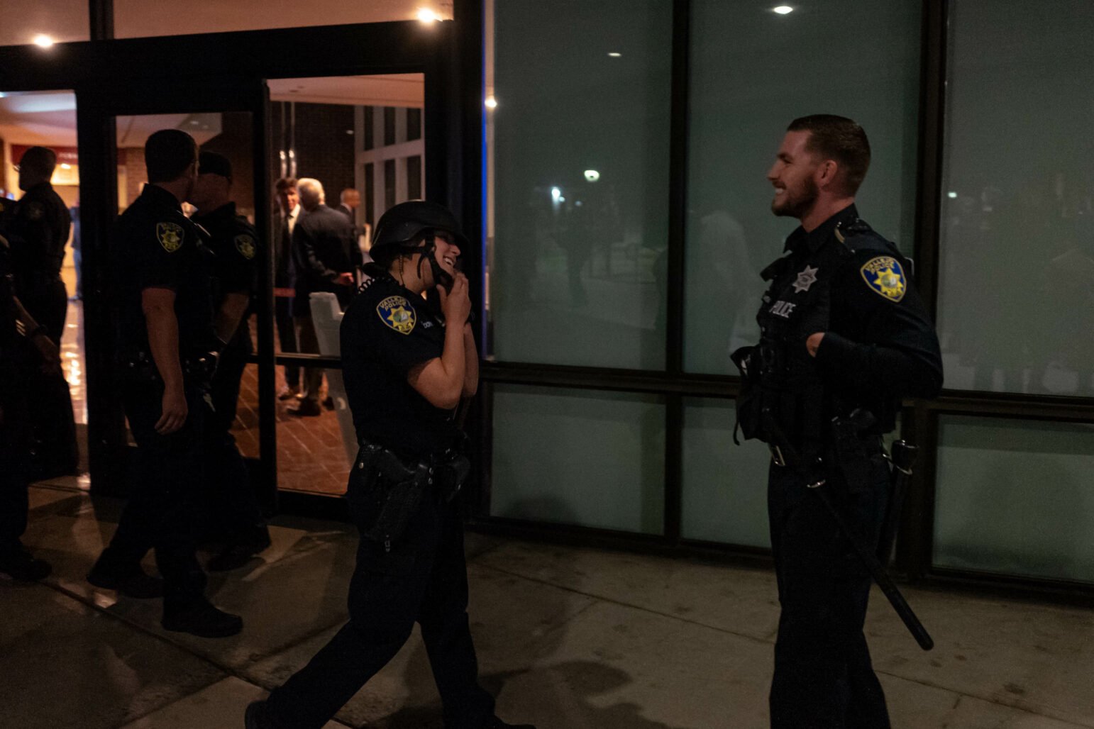Two police officers in conversing outside a building at night. The officer on the left is a woman and is taking off a tactical helmet. The officer on the right is a man. Both are smiling broadly. Other officers and people are visible in the background.
