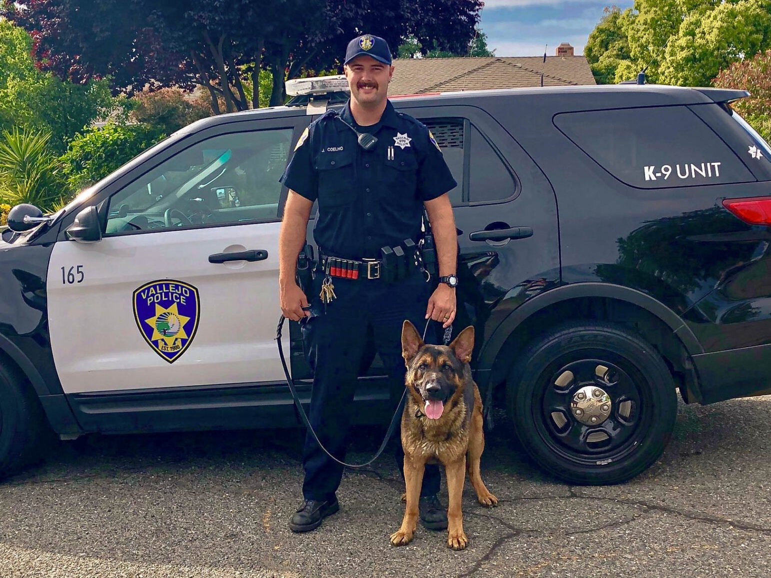 A police officer in uniform and a K-9 unit dog posing next to a police SUV. The officer, smiling and standing confidently, holds the leash of the German Shepherd, which sits beside him looking directly at the camera. The SUV has the Vallejo Police emblem and "K-9 UNIT" marked on it, set against a residential background with lush greenery.