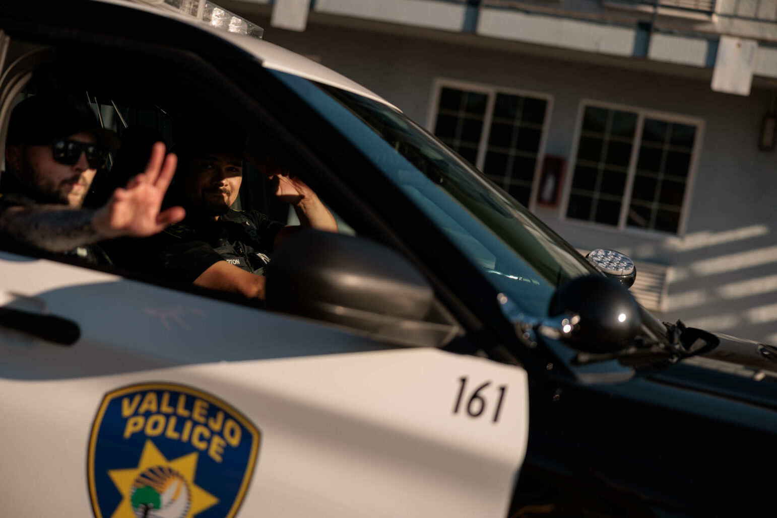 Two police officers in a patrol SUV, visible through the passenger side window, with one officer waving and the other raising his hand in a greeting or gesture. The car, marked with 'Vallejo Police' and the unit number '161', is partially shaded, with a motel partially visible in the background.