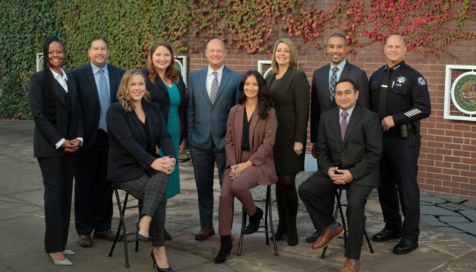 A group of 10 professionals in business attire posing for a photo outdoors. The setting features a backdrop of a brick wall covered in ivy. The group includes five women and five men, one of whom is dressed in a police uniform. They are smiling and standing or seated in two rows, projecting a collaborative and formal team image.