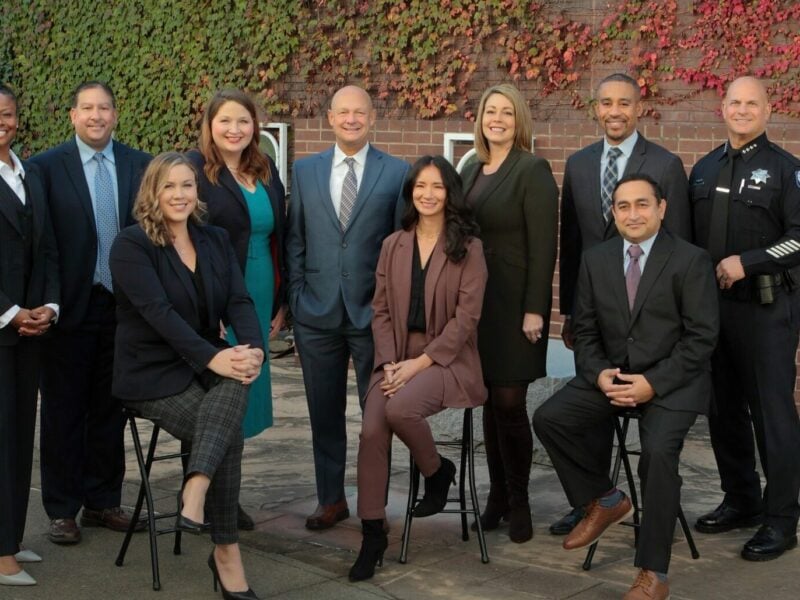 A group of 10 professionals in business attire posing for a photo outdoors. The setting features a backdrop of a brick wall covered in ivy. The group includes five women and five men, one of whom is dressed in a police uniform. They are smiling and standing or seated in two rows, projecting a collaborative and formal team image.