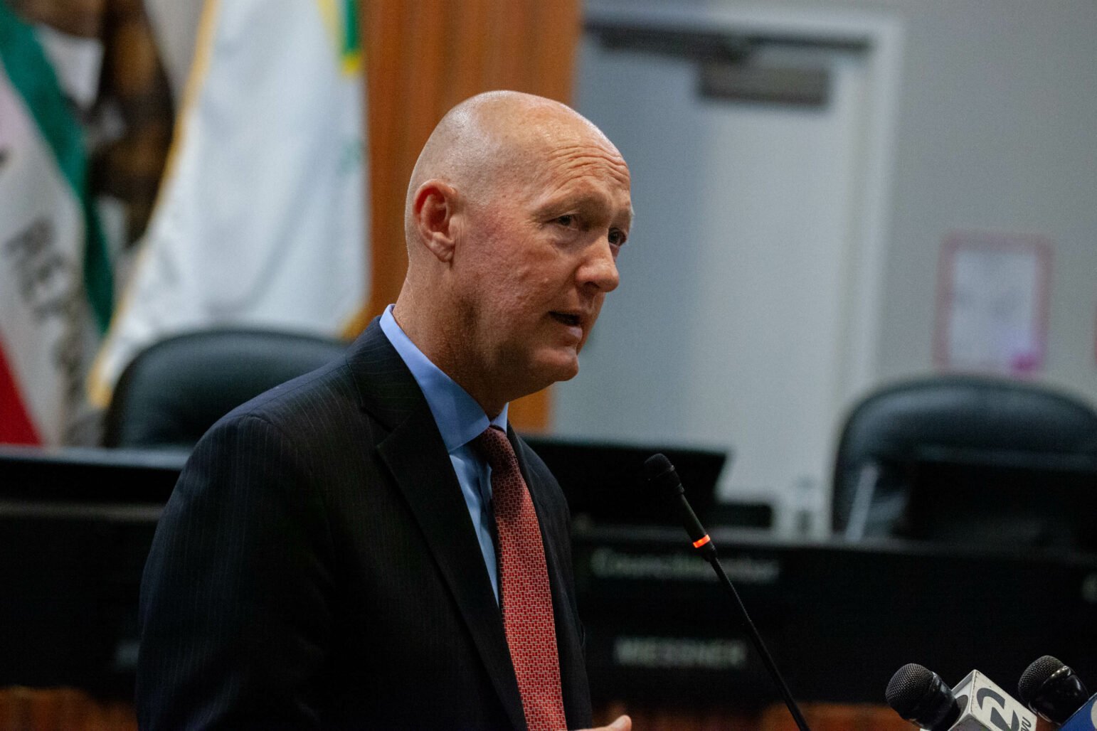 A man in a suit, identified as Greg Nyhoff, is speaking at a podium with a microphone. He appears to be addressing an audience in a city council chambers. Behind him, there are chairs and a California state flag is partially visible.