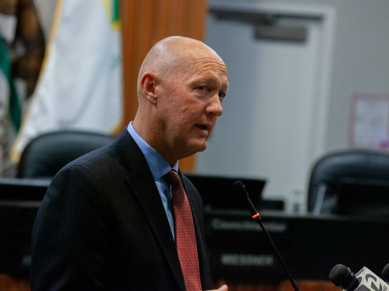 A man in a suit, identified as Greg Nyhoff, is speaking at a podium with a microphone. He appears to be addressing an audience in a city council chambers. Behind him, there are chairs and a California state flag is partially visible.