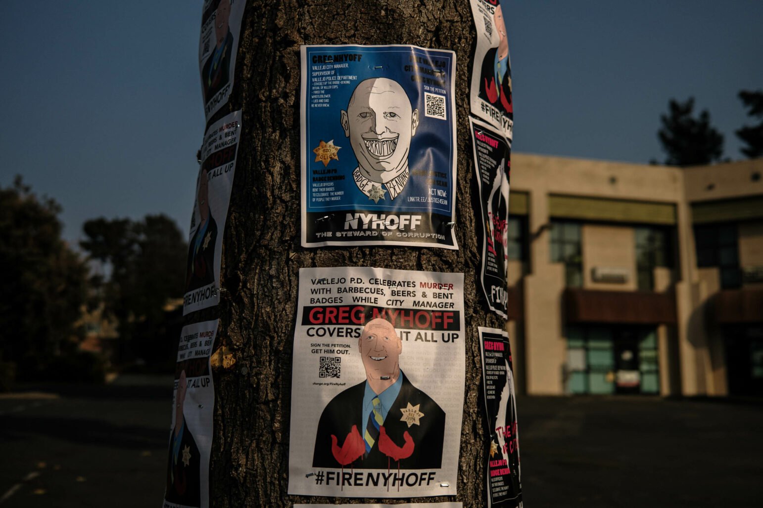 A tree covered in posters criticizing Greg Nyhoff, the former Vallejo City Manager, is shown. The posters depict Nyhoff with captions accusing him of covering up police misconduct and corruption. The background includes a building and a parking lot, indicating a public area.