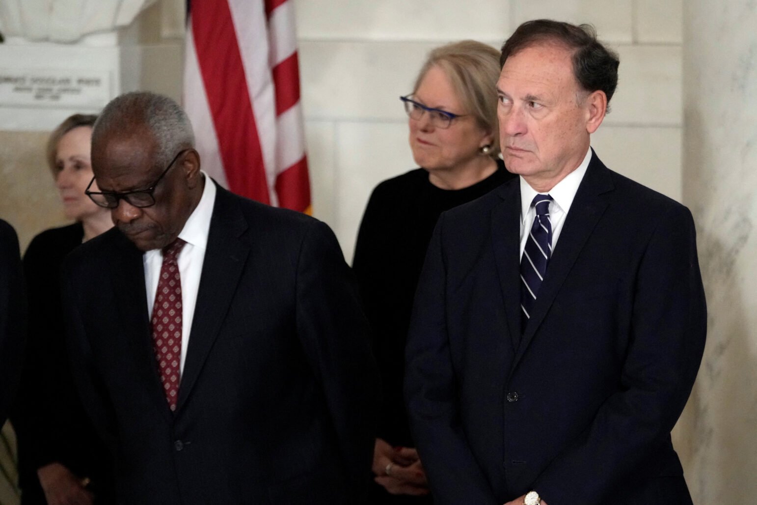 U.S. Supreme Court Justices Clarence Thomas and Samuel Alito standing together in a formal setting. Justice Thomas is looking down, while Justice Alito is looking ahead. Both are dressed in dark suits, and an American flag is visible in the background.