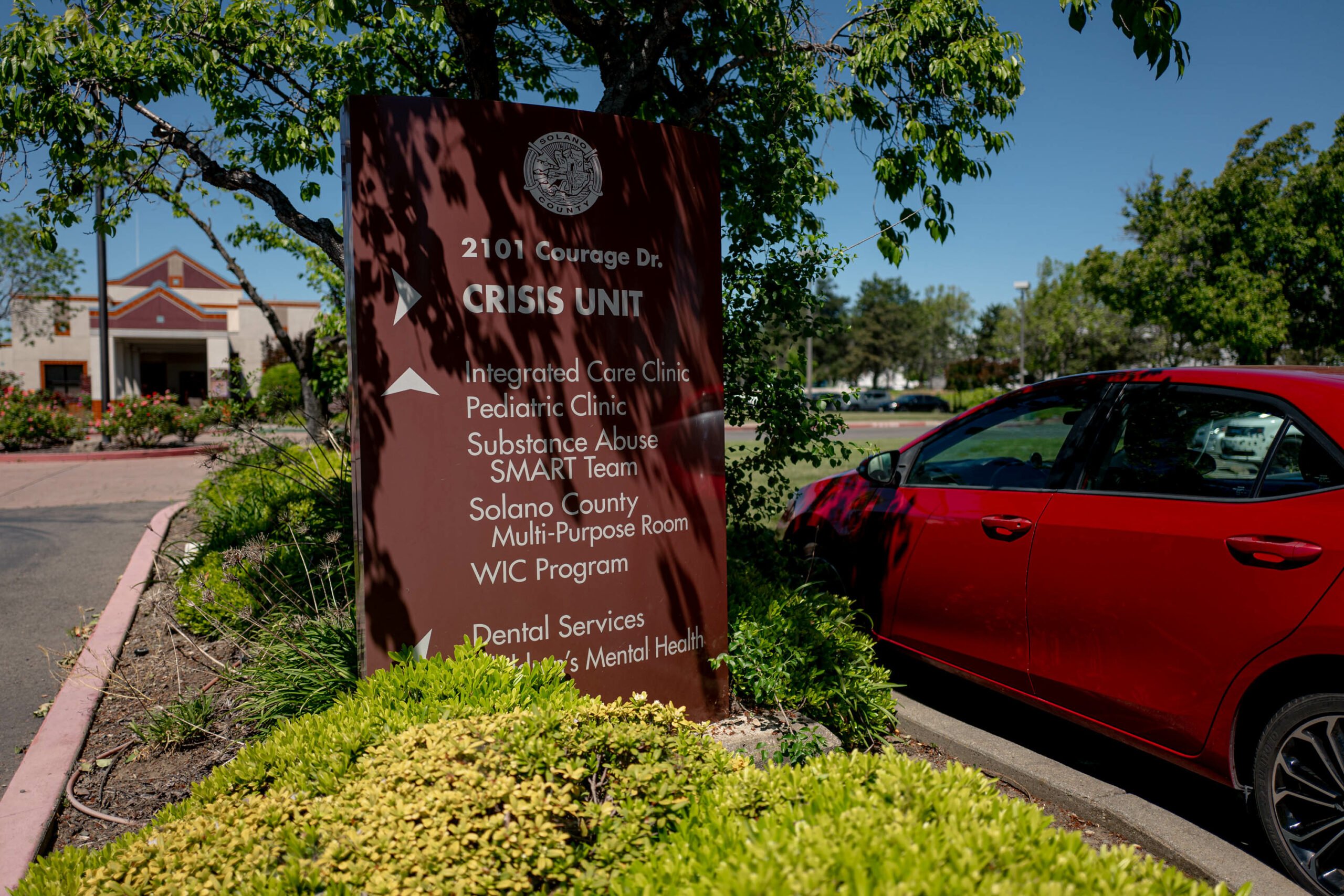 An outdoor view of a healthcare facility's entrance sign, labeled 'CRISIS UNIT' at 2101 Courage Dr. The sign lists various services such as Integrated Care Clinic, Pediatric Clinic, Substance Abuse, SMART Team, and more. The backdrop includes a modern building, lush greenery, and a parked red car, set under a clear blue sky.
