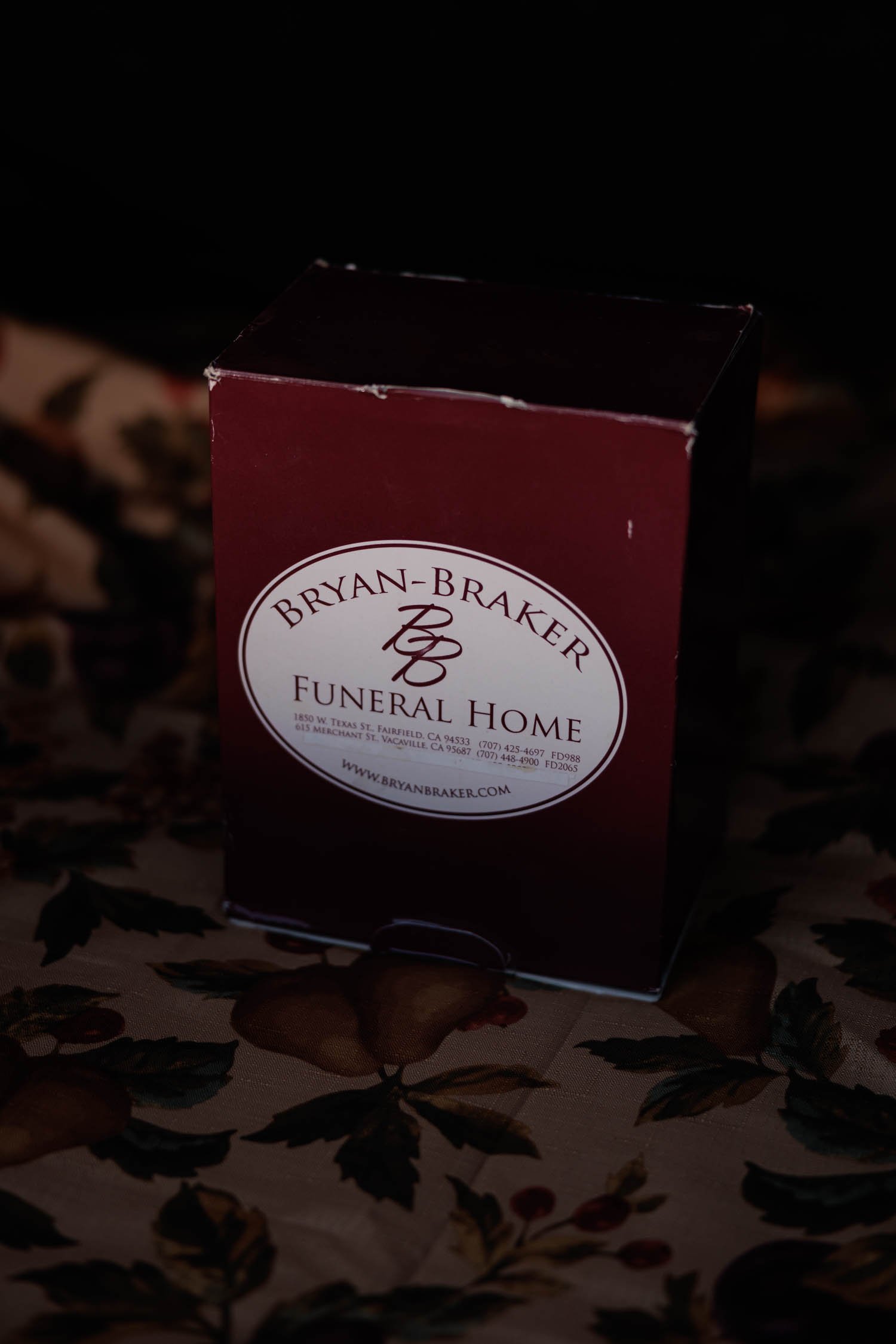 Close-up of a dark red box with the logo of Bryan-Braker Funeral Home printed in white. The box is slightly worn at the edges and sits against a dark, floral-patterned backdrop, giving it a somber and dignified appearance.
