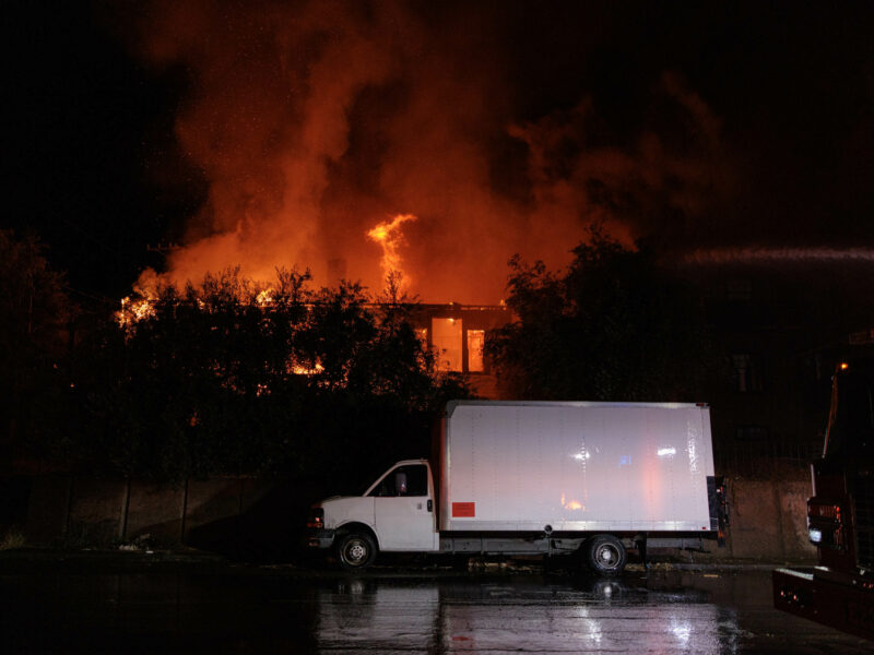 A white box truck parked on the street in front of a burning building at night. Flames and smoke are rising high into the sky from the roof of the building.