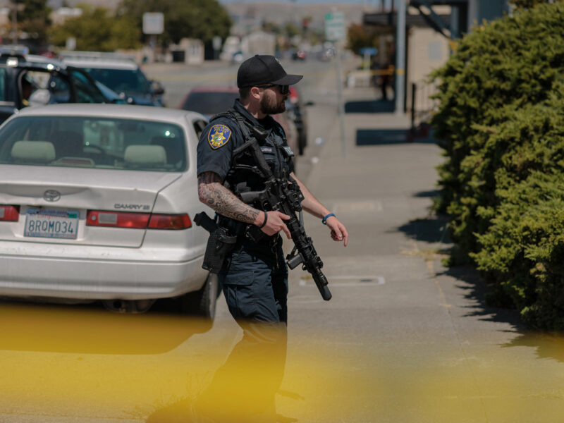 A police officer holding a rifle approaches a residence during daytime.