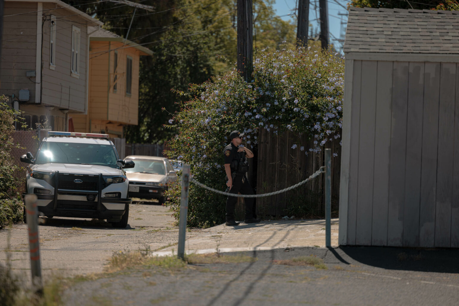 A sheriff's deputy stands in an alleyway near a wooden fence covered with blooming flowers during daylight. He holds a handgun pointed at the ground in his right hand.