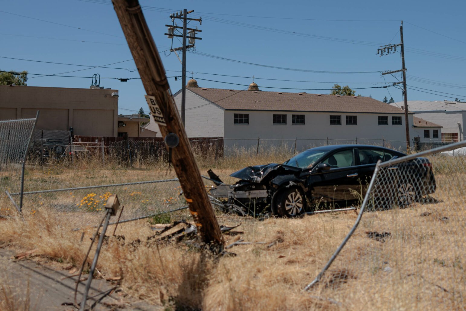 A car accident scene under a clear blue sky. A black sedan with significant front-end damage is situated off-road, having collided with a leaning wooden utility pole. Debris from the car and pole are scattered around the dry, grassy area, which is bordered by a chain-link fence. 