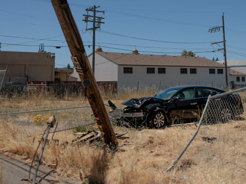 A car accident scene under a clear blue sky. A black sedan with significant front-end damage is situated off-road, having collided with a leaning wooden utility pole. Debris from the car and pole are scattered around the dry, grassy area, which is bordered by a chain-link fence.