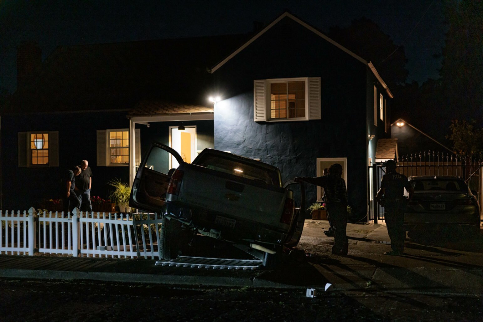 A blue house at night with a silver pickup truck crashed into the white picket fence in front. The truck's front is elevated and appears to be lodged on the fence. Several people, including police officers, are standing near the house, and the area is lit by a porch light and police car lights.