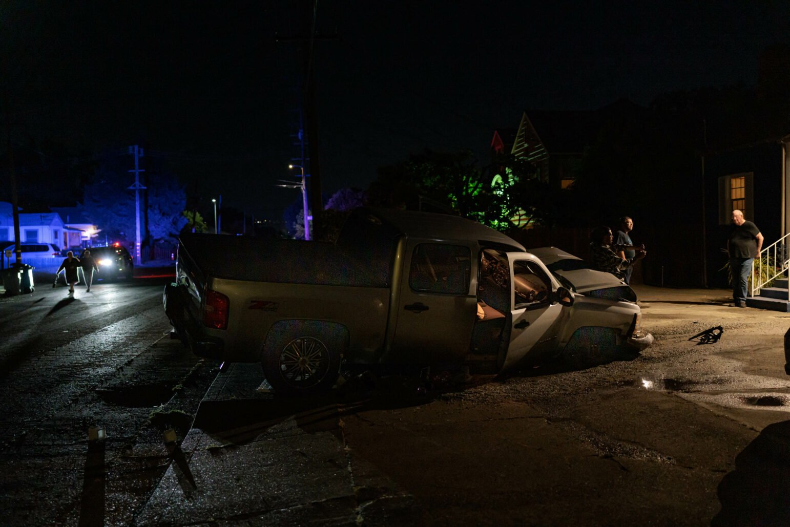 Nighttime view of a silver pickup truck that has crashed into a white picket fence in front of a dark blue house. A police officer is inspecting the truck, and another person is sitting inside the vehicle. The house is partially illuminated by its porch light and nearby police car lights.