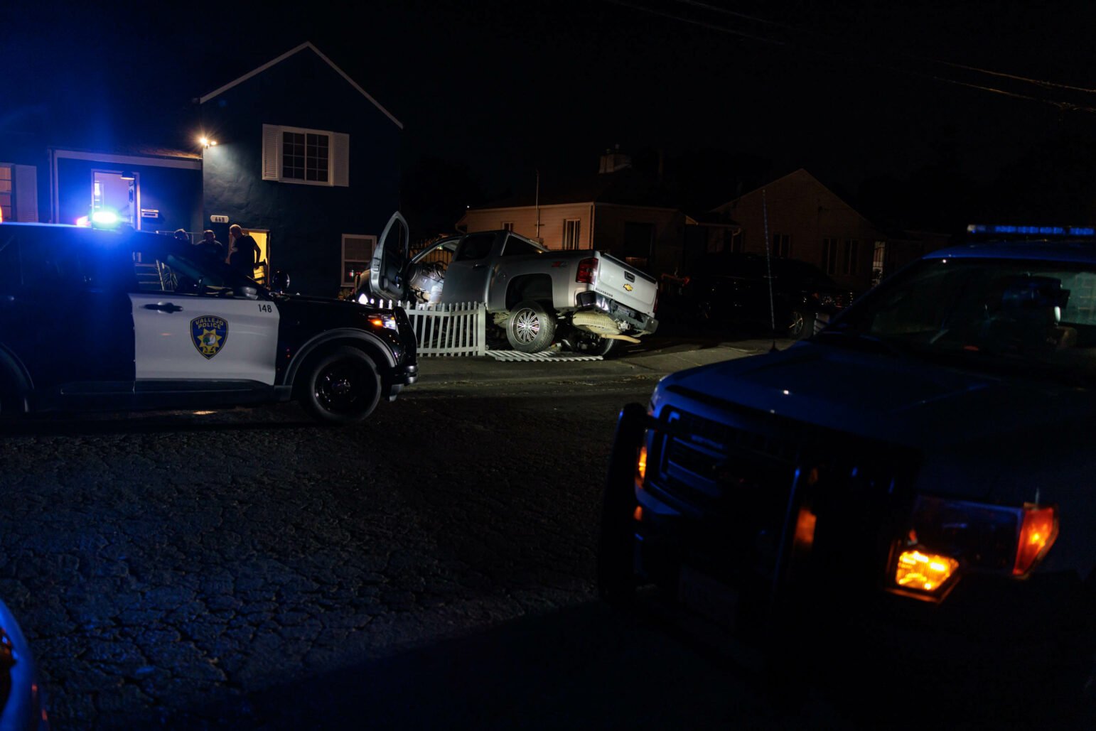 Nighttime scene showing a silver pickup truck that has crashed into a white picket fence in front of a dark blue house. Two police cars with flashing lights are parked nearby, and several officers are standing around the scene.