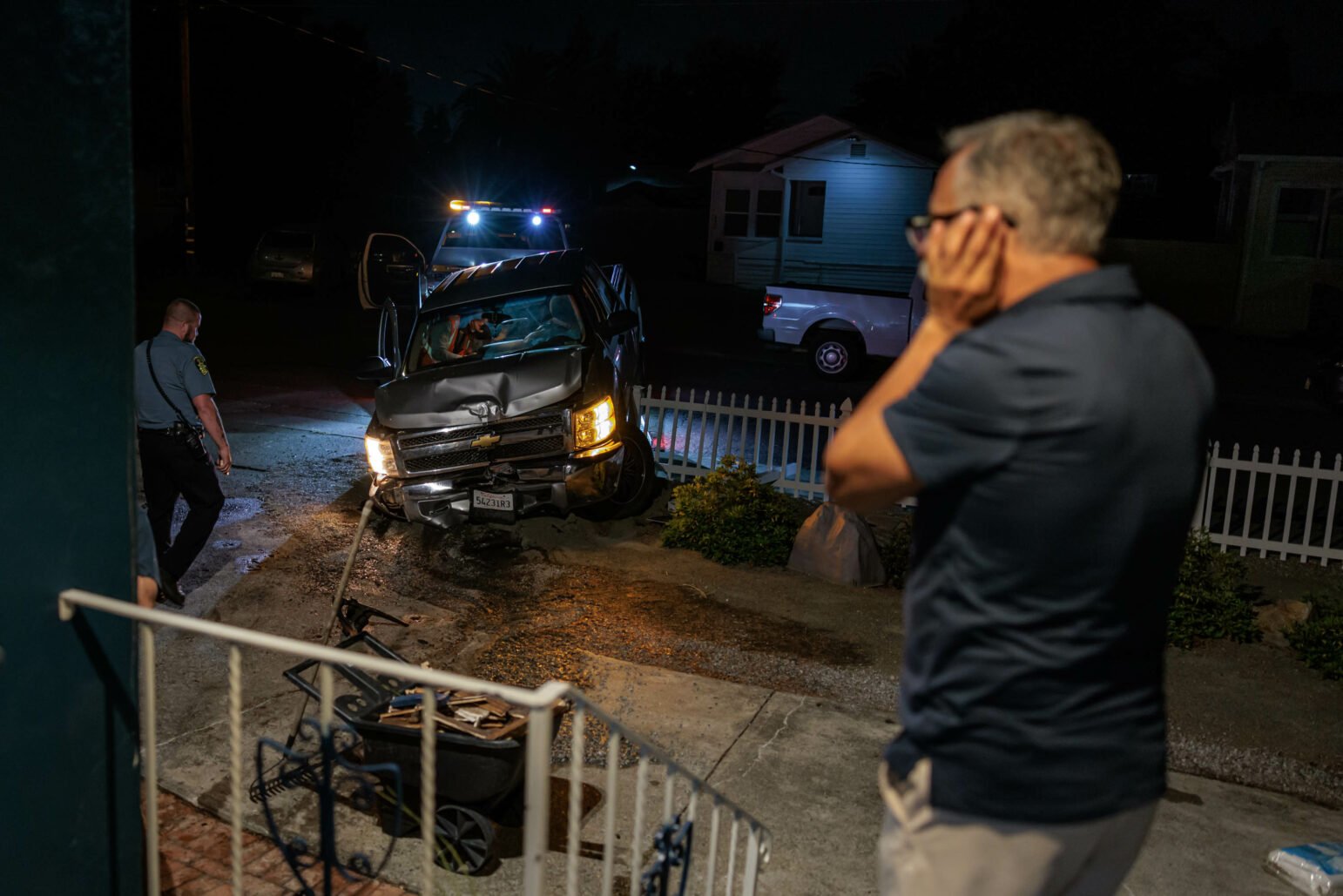 Close-up nighttime scene of a damaged silver pickup truck crashed into a white picket fence. A police officer is inspecting the scene while a man in a blue shirt stands nearby while speaking on the phone. The area is lit by the flashing lights of a tow truck and police car.