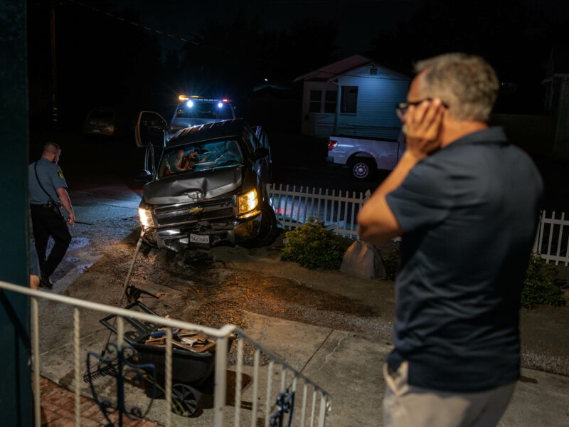 Close-up nighttime scene of a damaged silver pickup truck crashed into a white picket fence. A police officer is inspecting the scene while a man in a blue shirt stands nearby while speaking on the phone. The area is lit by the flashing lights of a tow truck and police car.