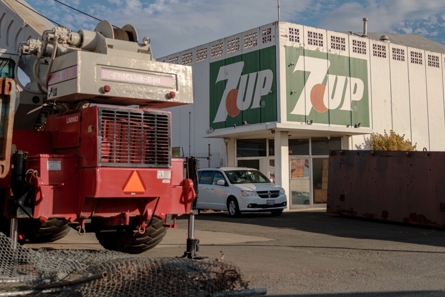 A former 7Up factory building with a large logo on the side, viewed from the parking lot. A white van is parked in front, and an industrial crane is partially visible on the left. The scene is lit by the late afternoon sun.