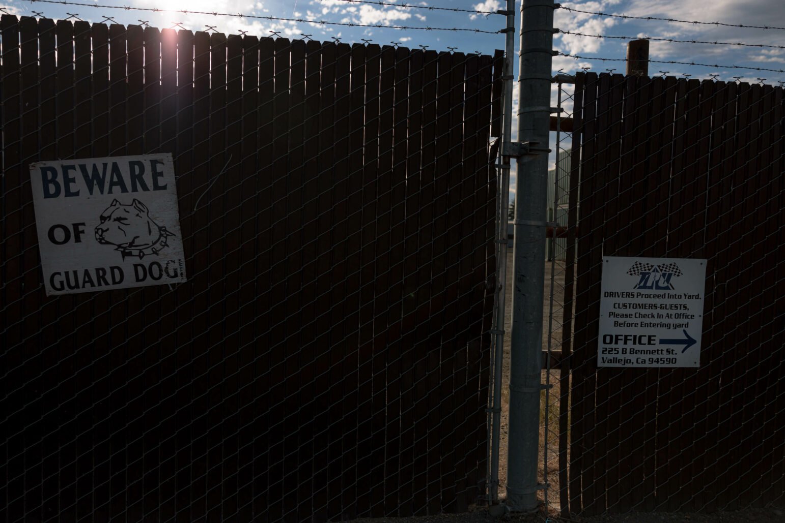 A chain-link fence topped with barbed wire, featuring a 'Beware of Guard Dog' sign on the left and an instruction sign for drivers and customers on the right. The sky is partly cloudy with sunlight shining through, casting shadows on the fence.
