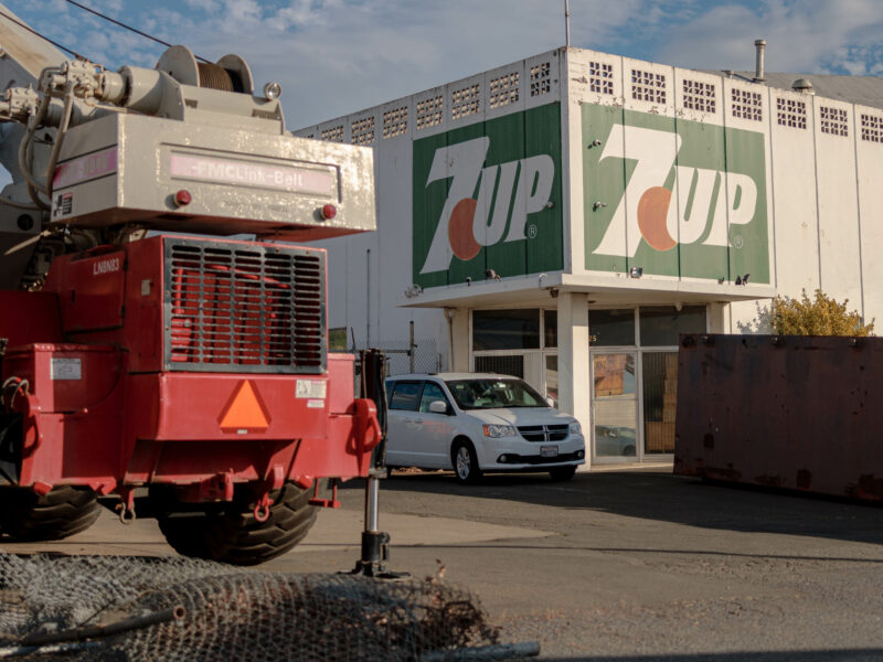 A former 7Up factory building with a large logo on the side, viewed from the parking lot. A white van is parked in front, and an industrial crane is partially visible on the left. The scene is lit by the late afternoon sun.