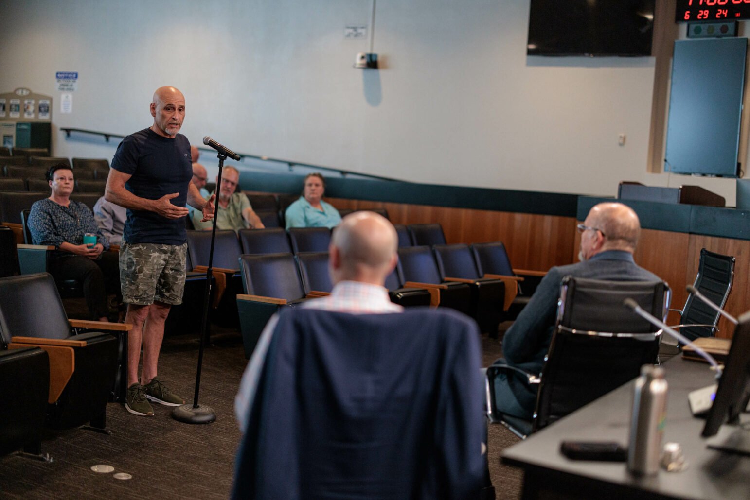A man in a dark shirt and camouflage shorts stands at a microphone speaking to an audience during a town hall meeting. Seated behind him are several attendees, including a woman in a blue patterned shirt and a man in a green shirt.