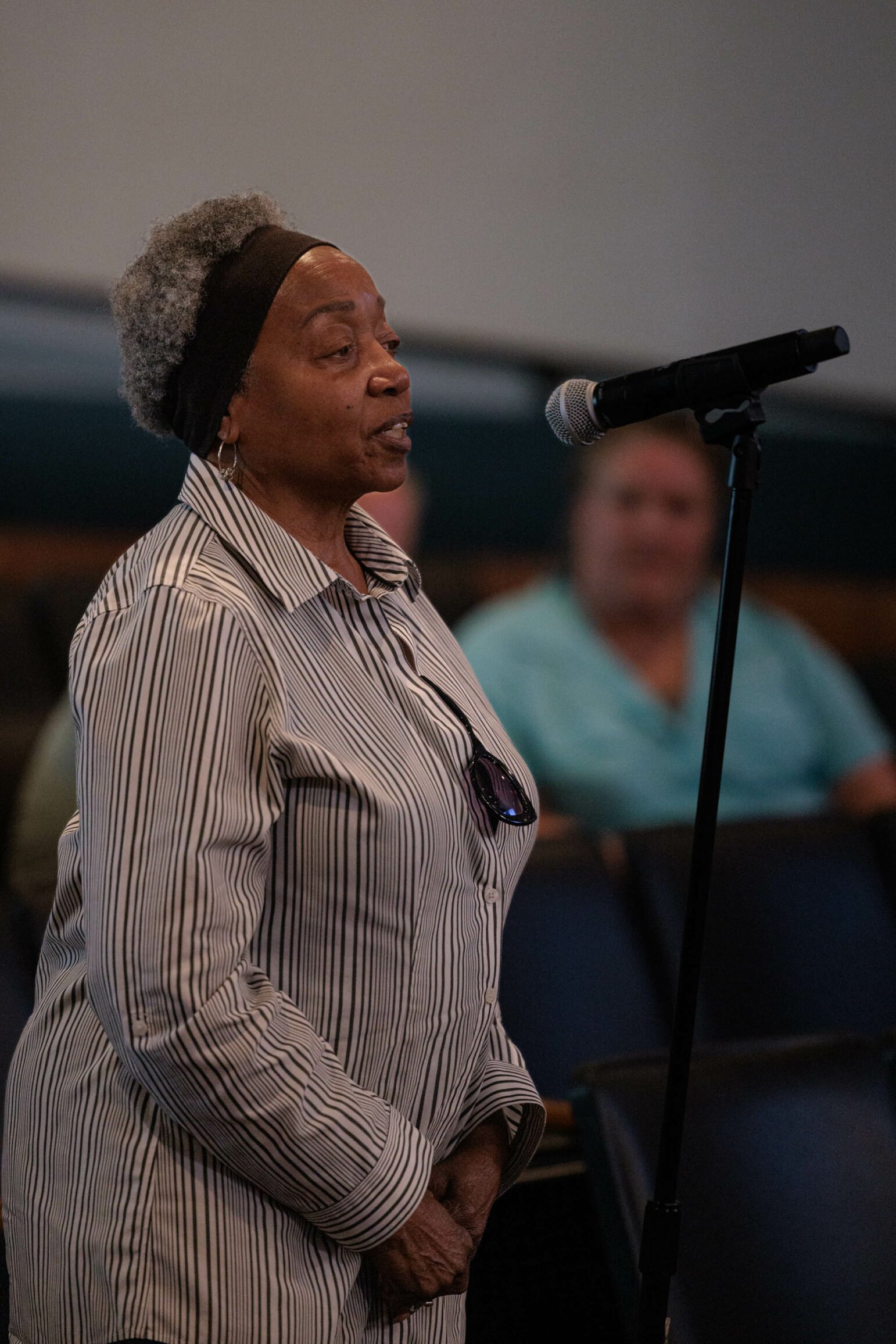 An older woman with gray hair and a headband speaks into a microphone during a town hall meeting. She is wearing a striped shirt and sunglasses hanging from her shirt. Attendees are seated in the background, listening attentively.