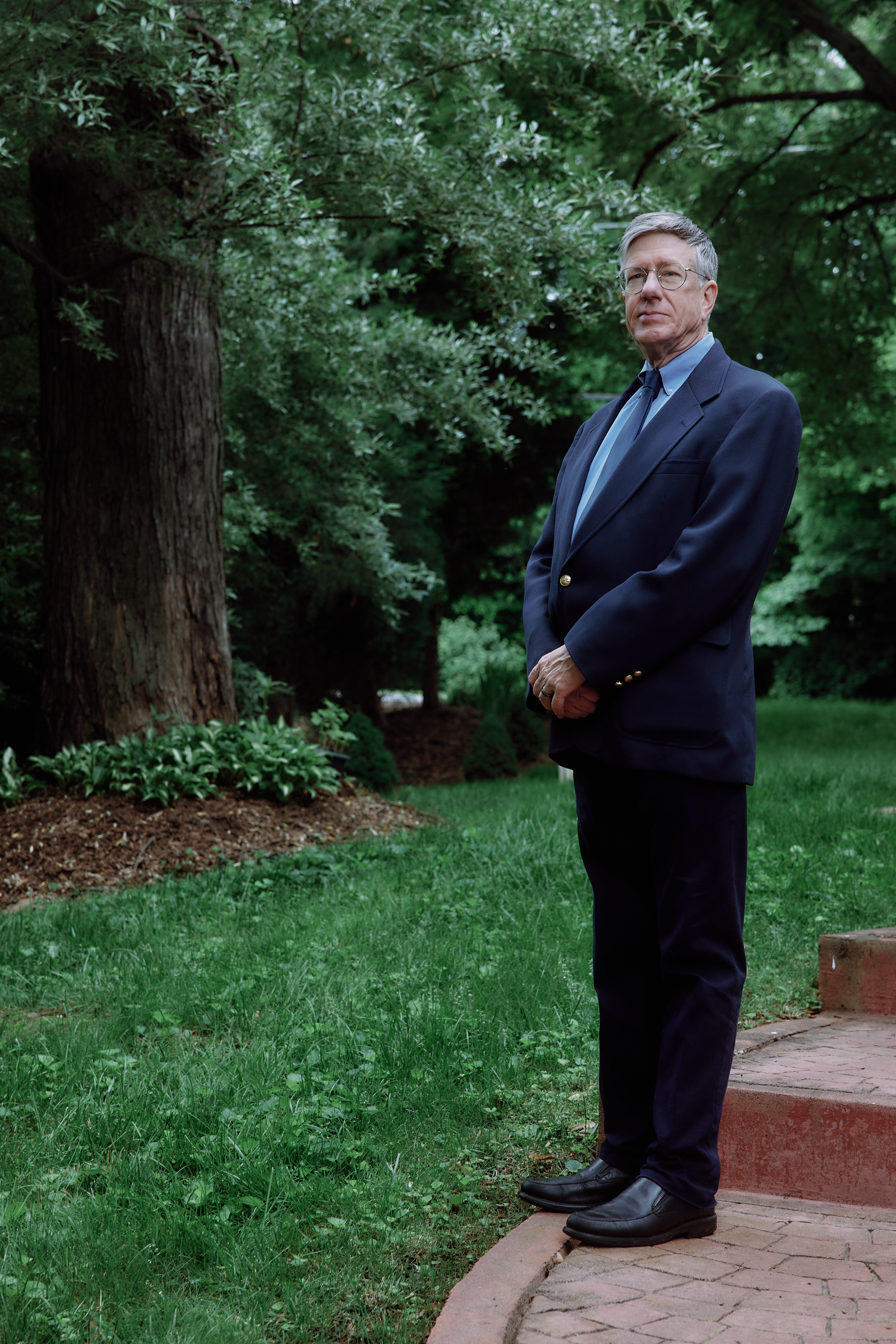 Portrait of a doctor standing outdoors in a serene garden setting. He is dressed in a formal navy blue blazer with gold buttons, a light blue shirt, a dark blue tie, and dark trousers. He looks directly at the camera with a composed expression, his hands clasped in front of him. Large trees and lush greenery provide a natural backdrop, enhancing the dignified atmosphere of the scene.