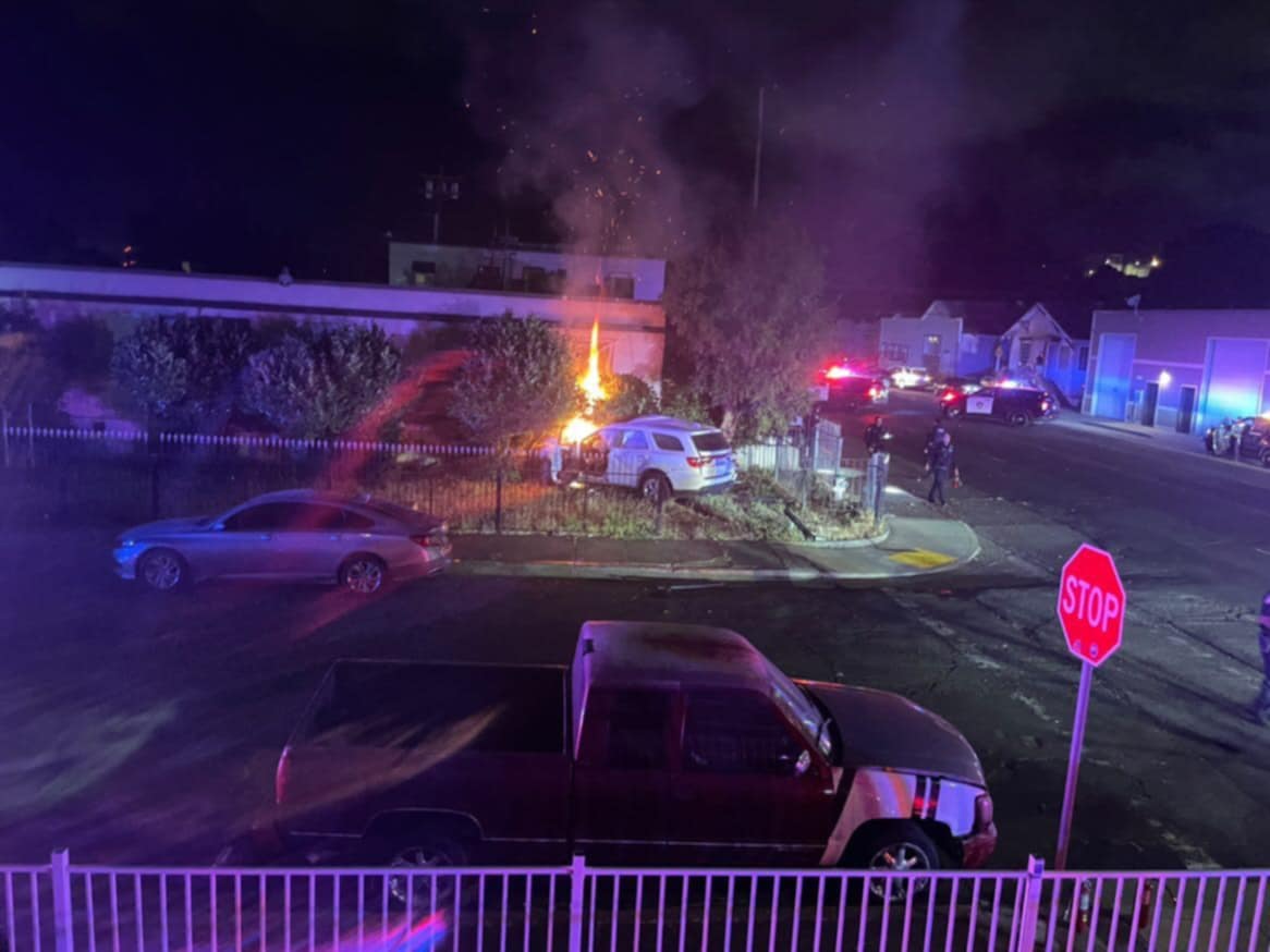 A nighttime scene of a car crash with a white SUV on fire on a grassy area beside a road. The SUV has crashed into a building on the corner. Police cars with flashing lights are parked nearby, and several officers are on the scene. Other vehicles and houses are visible in the background, with a stop sign in the foreground.