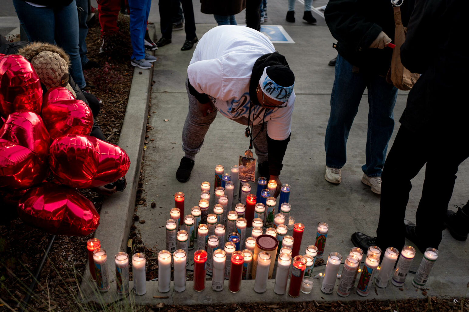 A group of people gathers around a memorial for Willie McCoy. A person in the center, wearing a white T-shirt and a headband, bends down to light a candle among many already lit candles arranged on the ground. Red heart-shaped balloons are on the left side of the image.