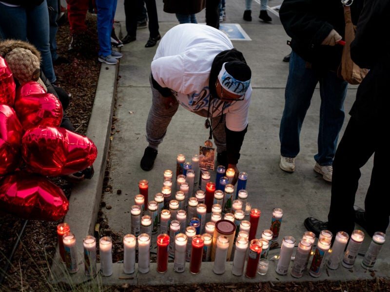 A group of people gathers around a memorial for Willie McCoy. A person in the center, wearing a white T-shirt and a headband, bends down to light a candle among many already lit candles arranged on the ground. Red heart-shaped balloons are on the left side of the image.
